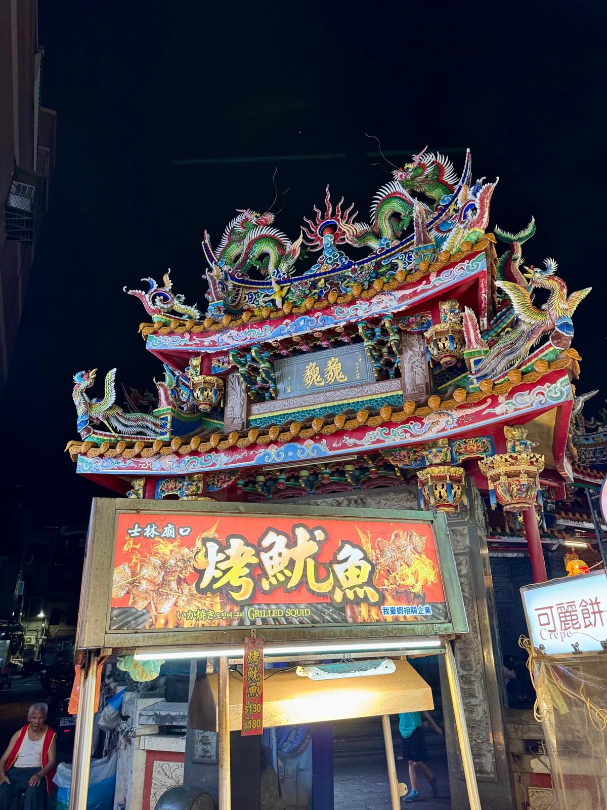 Night Market Temple at night with man sitting and grilled squid stall sign under it