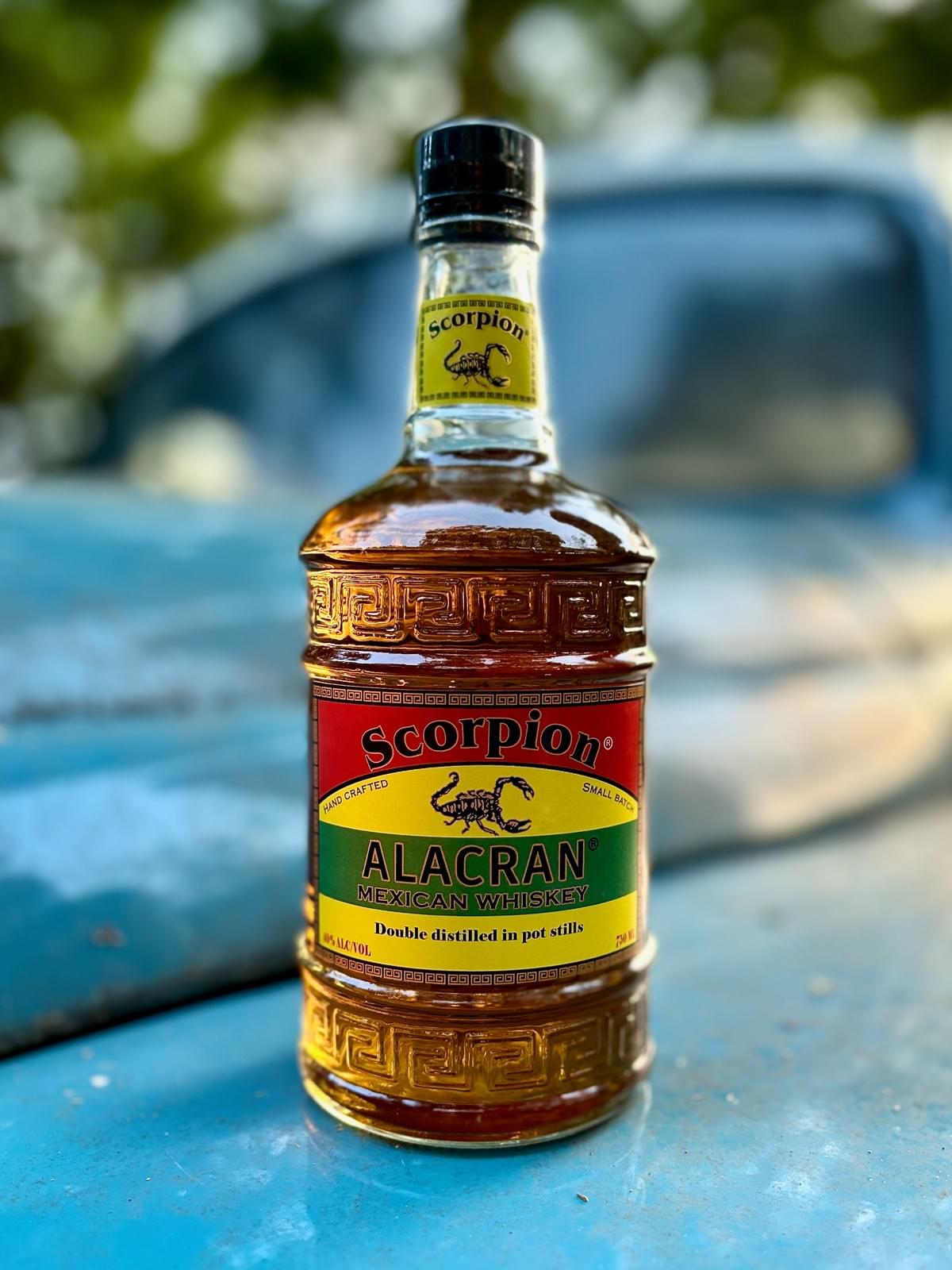 750mL bottle of ALACRAN Mexican Corn Whiskey, atop the hood of a turquoise 1940s truck.