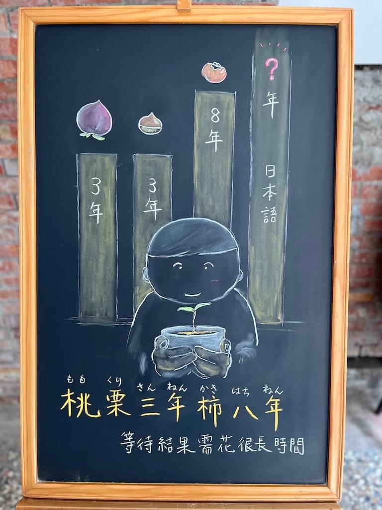 小さい黒板にチョークで絵が描いてあります。日本のことわざを紹介するための絵です。下部に横書きで「桃栗三年柿八年」と書いてあります。中央に白い小ぶりの植木鉢を両手で前に持った男の子のイラストが書いてあります。鉢からは小さな芽が出たところです。背景に棒グラフがあり、３年、３年、８年の長さで縦に伸びています。それらの上に桃、栗、柿のイラストがあります。一番右に「？年」と書いたグラフがあり、「日本語」と書いてあります。