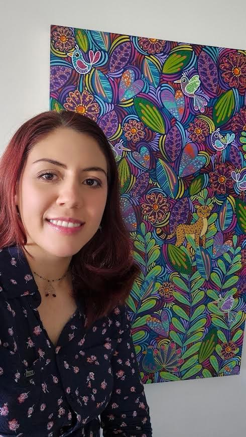 Colombian artist Angela María Medina Olano (AMMO) standing beside one of her colorful nature inspired artworks featuring birds, flowers, leaves, and intricate organic patterns.