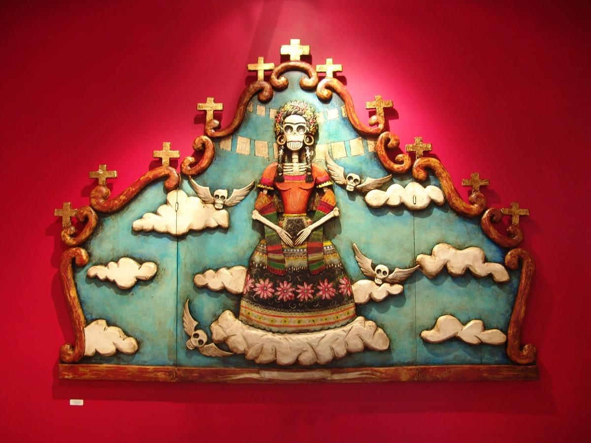 Day of the Dead inspired folk art by Gennaro Garcia featuring a female skeleton figure surrounded by clouds, crosses, and winged skulls against a vibrant red background with traditional Mexican symbolism