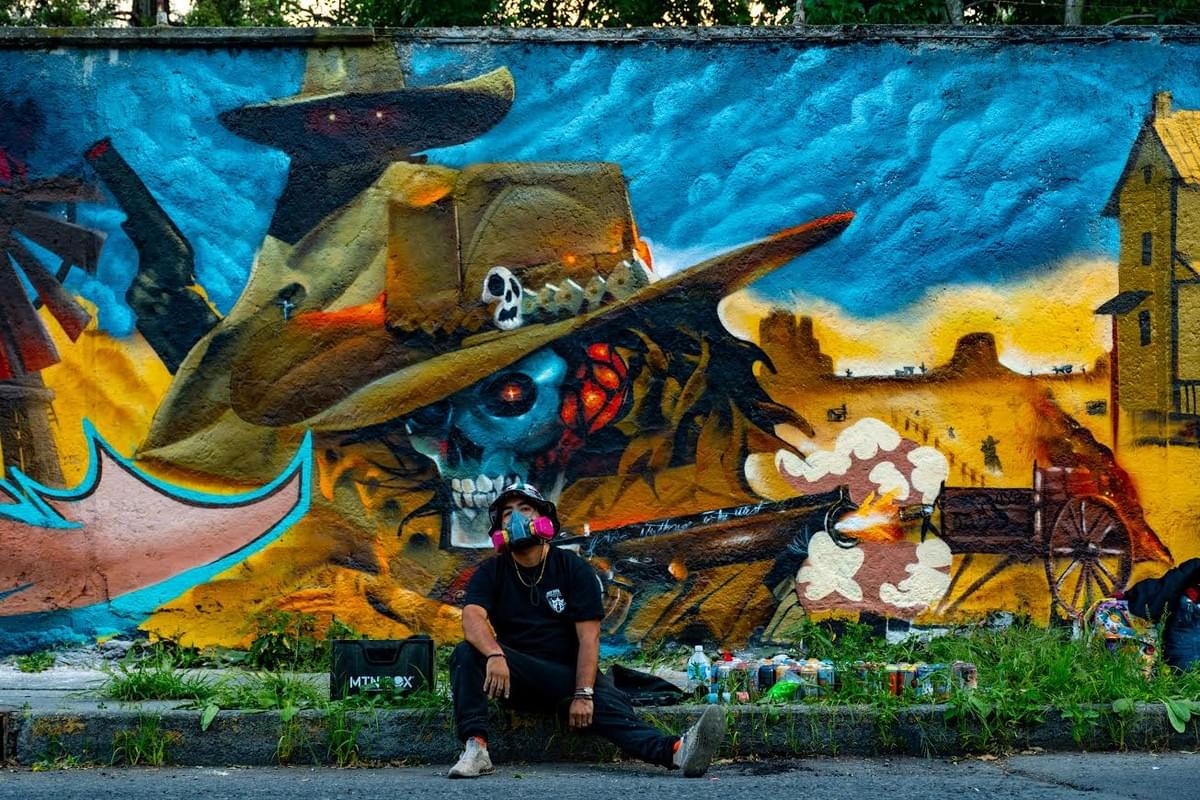 Large scale mural by Querétaro artist Magia Negra featuring a skeletal cowboy figure in a wide brim hat surrounded by desert imagery, glowing eyes, dramatic blue skies, and western inspired fantasy symbolism while the artist sits in front of the wall with spray paint cans.