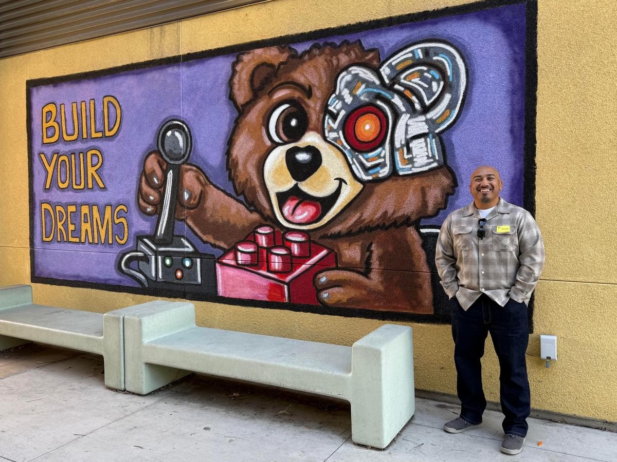 Colorful mural by Bad Hombre featuring a cartoon bear with a robotic eye and the phrase "Build Your Dreams" painted on a public wall