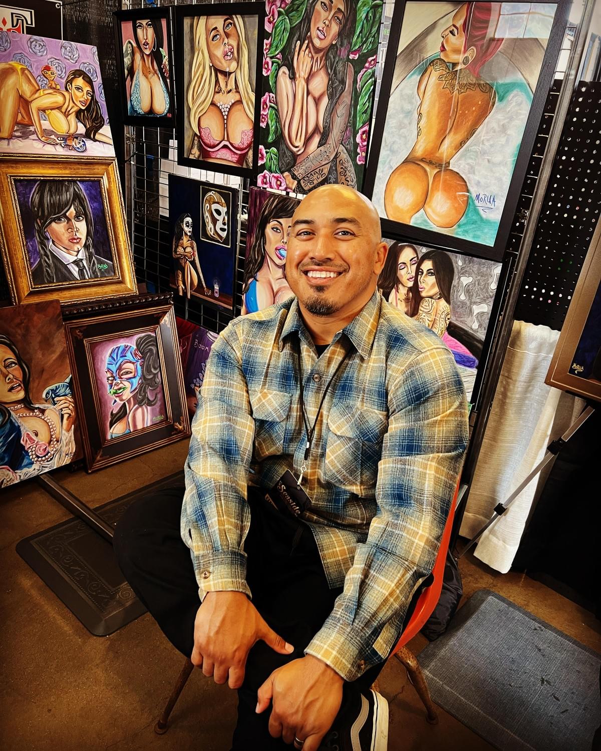 Artist Bad Hombre seated at an art booth surrounded by bold, colorful Chicano pop art paintings featuring portraits and cultural themes