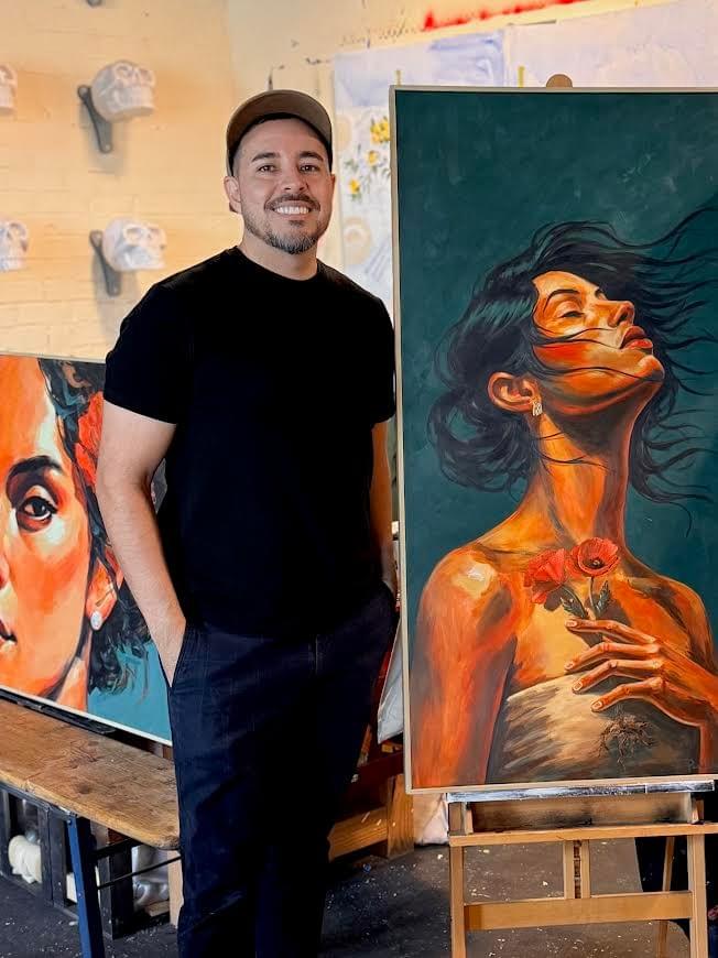 Phoenix artist Michael Padilla standing inside his studio beside a large contemporary portrait painting featuring a woman holding orange poppy flowers, surrounded by expressive figurative artwork and warm natural light.