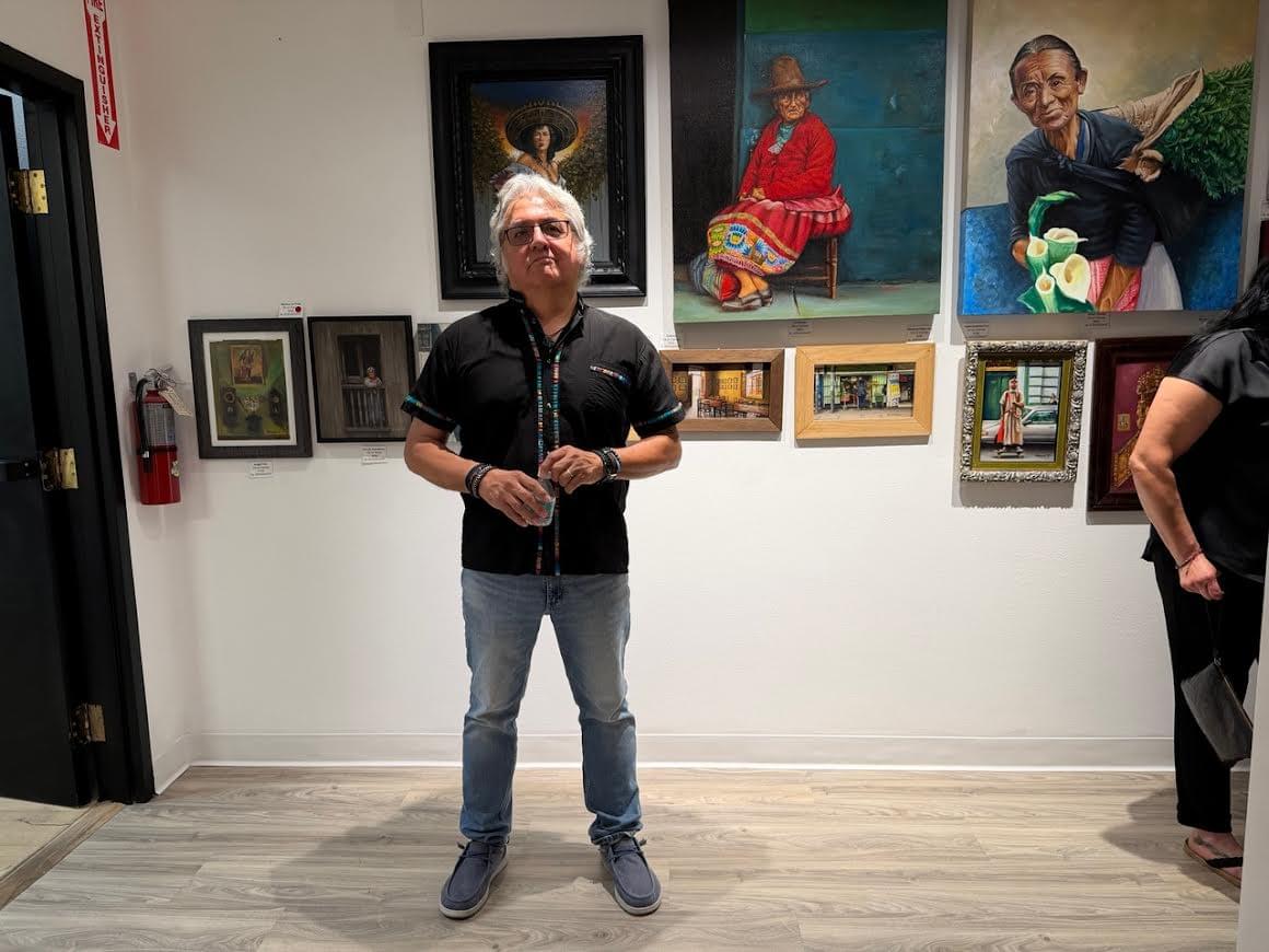 Artist Roger Triviños standing in a gallery exhibition surrounded by paintings inspired by Peruvian culture, Latin American traditions, and everyday life.