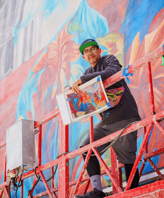Dominican mural artist Angurria standing on scaffolding while painting a large colorful mural, holding artwork in progress