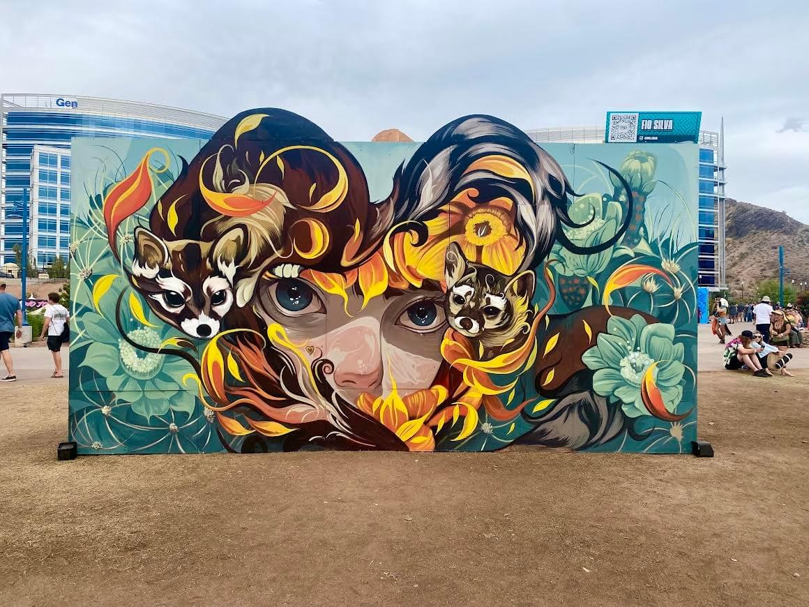Portrait mural by Fio Silva featuring a female face surrounded by flowing organic shapes, animals, and vibrant natural elements in a public outdoor setting