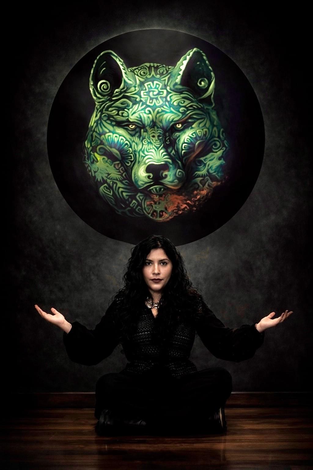 Portrait of Mexican artist Yuma Díaz seated beneath a glowing symbolic wolf artwork inspired by Indigenous mythology, ancestral imagery, and contemporary Mexican visual storytelling.