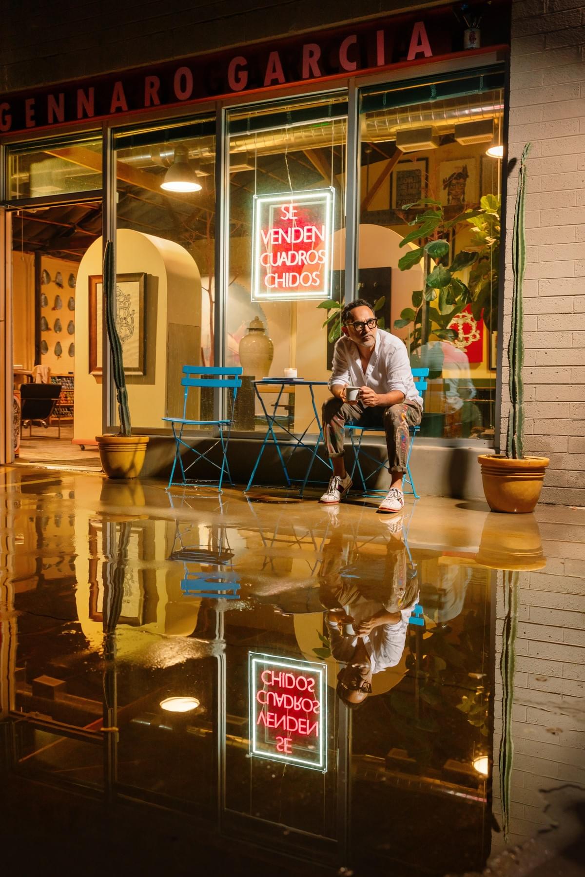 Artist Gennaro Garcia sitting outside his Phoenix art gallery and studio at night with neon signage, colorful reflections, and Mexican inspired artwork displayed inside the space