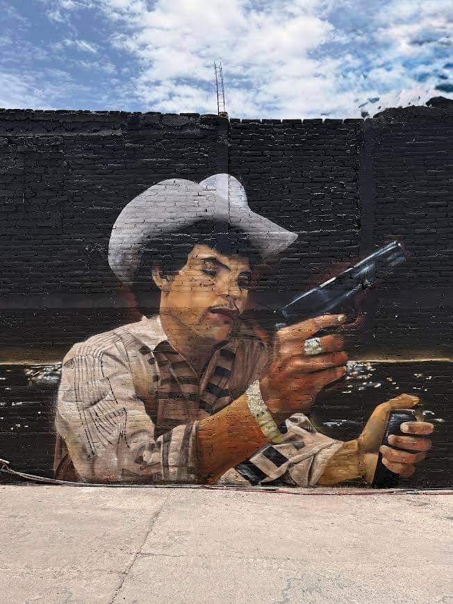 Urban mural by Mexican artist Magia Negra featuring a cowboy holding a spray paint can and paint sprayer, blending street culture, realism, and graffiti inspired imagery against a dark brick wall in Mexico.