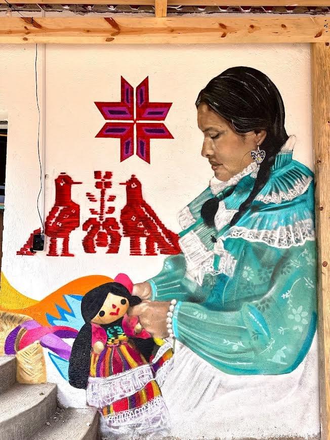 Mural by Mexican street artist Magia Negra featuring an Indigenous woman in traditional clothing holding a handmade doll, surrounded by colorful folk art inspired patterns and cultural symbolism in Mexico.