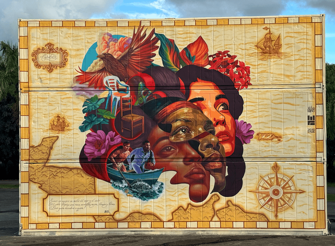 Color Caribe mural by Angurria featuring layered Caribbean portraits, cultural symbols, and historical elements on a large outdoor wall