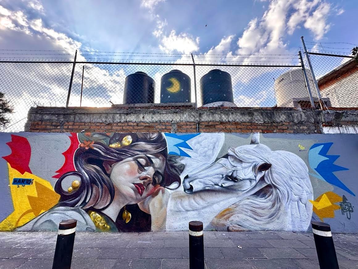 Street mural by Mexican artist Magia Negra featuring a woman resting beside a white horse, blending surreal fantasy imagery with soft realism and colorful urban mural elements in Querétaro, Mexico.