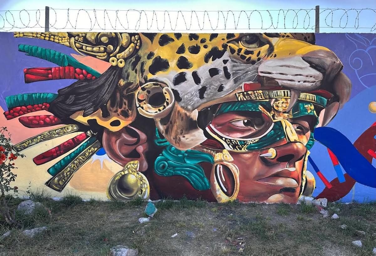 Colorful mural by Mexican street artist Magia Negra featuring a large Indigenous warrior portrait with ceremonial jewelry, jaguar symbolism, feathers, and bold Mesoamerican inspired imagery painted across an outdoor wall in Mexico.