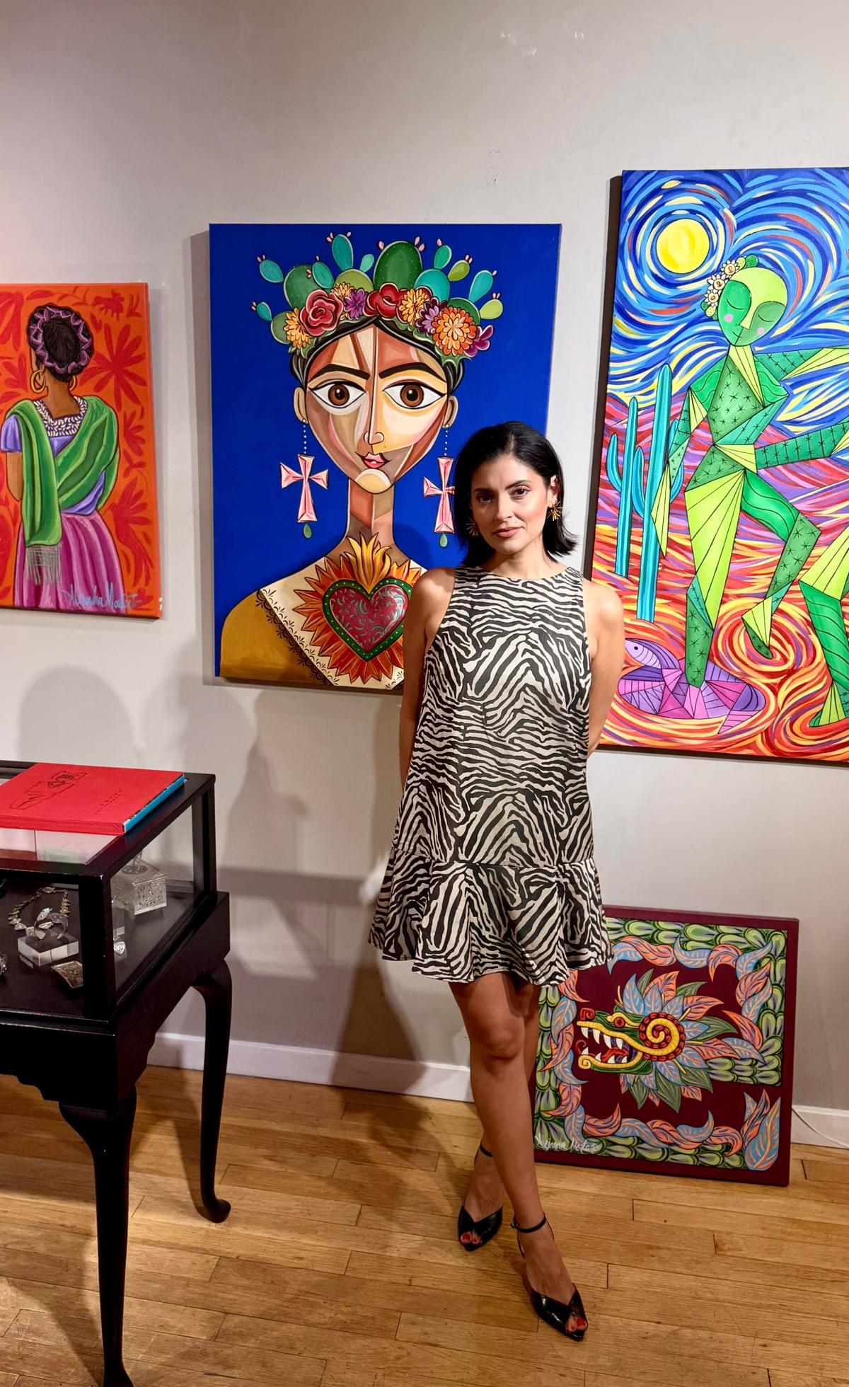 Alejandra Montaño standing beside her contemporary Mexican paintings inspired by culture, symbolism, and vibrant storytelling in her art studio.