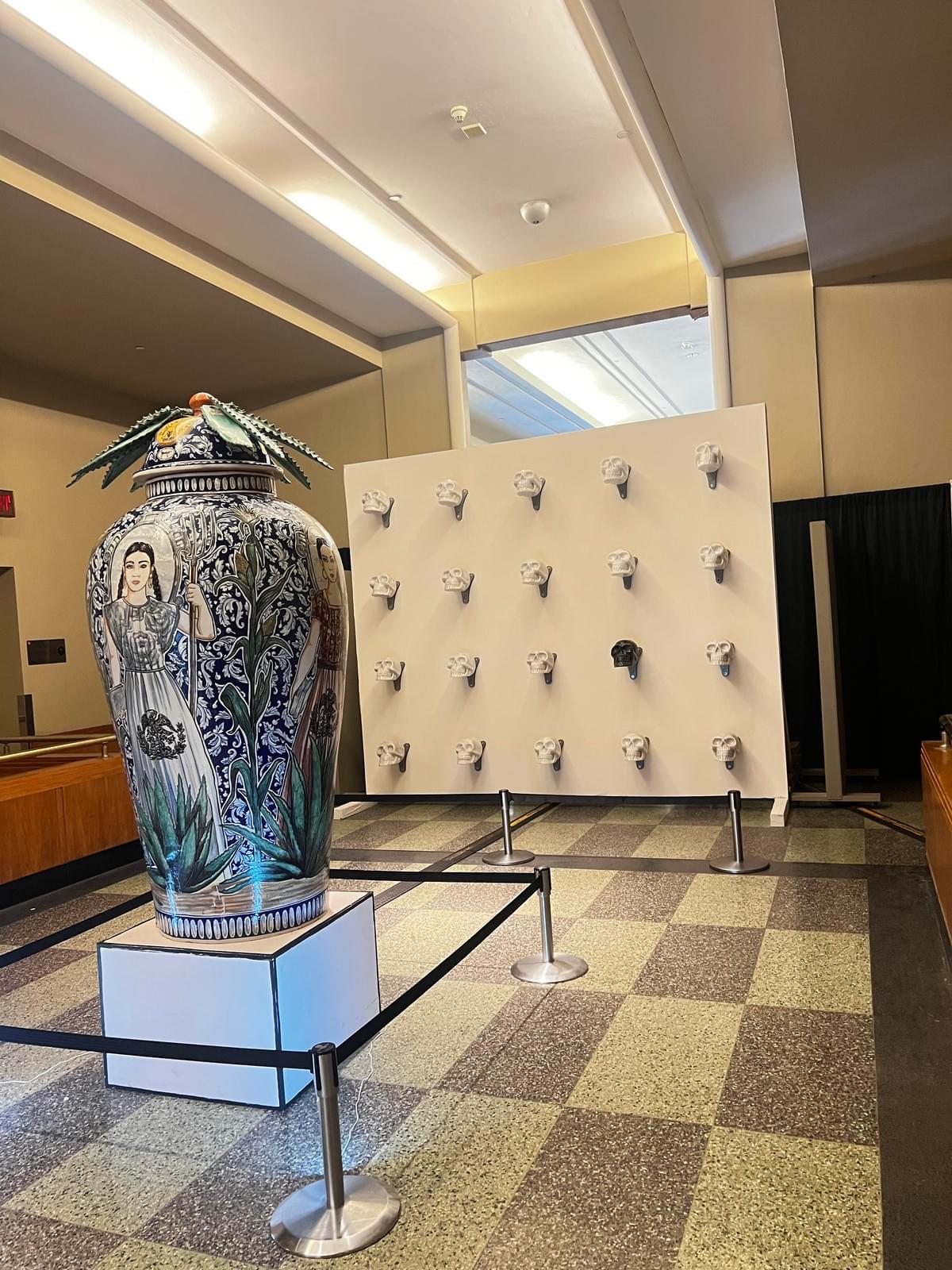Contemporary art installation by Gennaro Garcia featuring a large hand painted ceramic vessel with Mexican inspired figures and decorative skull sculptures displayed in a gallery setting