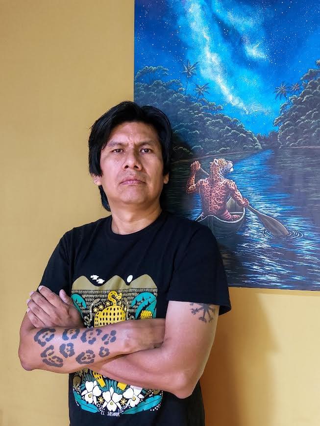 Peruvian artist César Chujutalli standing beside one of his Amazon-inspired paintings featuring a sacred jaguar canoeing beneath a star-filled rainforest sky.