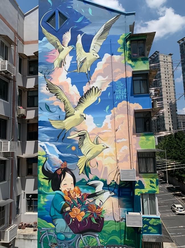 Large mural by Fio Silva featuring flying birds and a child riding a bicycle painted on a tall urban building facade