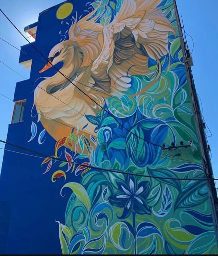 Large mural by Fio Silva featuring a swan in motion surrounded by flowing leaves and organic shapes on a tall city wall