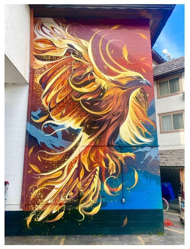 Large-scale mural by Fio Silva featuring a vibrant phoenix-like bird with flowing feathers in warm tones painted on a building exterior