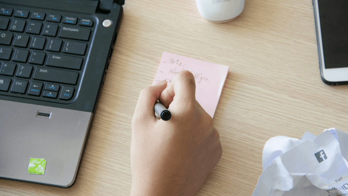 Editor writing a topic assignment on a sticky note beside a laptop