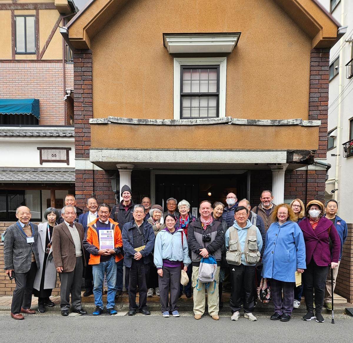 20+ YCAPS members and volunteers from the Yamamugi Incident Museum pose for a happy group photo in front of the museum. The museum is a small, ecclectic looking western style building attached to a more traditional Japanese house.