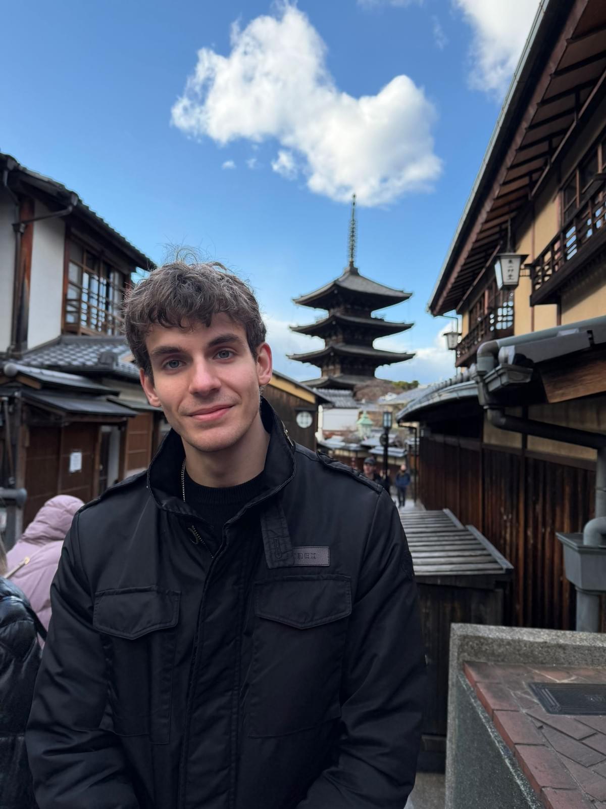 A fair skinned young man with wavy light hair looks at the camera with a light smile on his face. He is wearing a black medium-weight winter jacket while standing in a narrow, traditional looking Japanese street flanked by wooden buildings. In the background, a 5-story wooden padoda rises up into a blue sky.