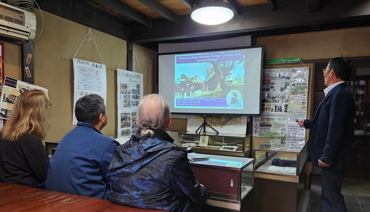 YCAPS members listen attentively to a presentation by a volunteer of the Namamugi Incident Museum. He points to a screen where a recolored photo of Namamugi Village is displayed. The building is old and Japanese styled, with informational posters and glass display cases scattered around.