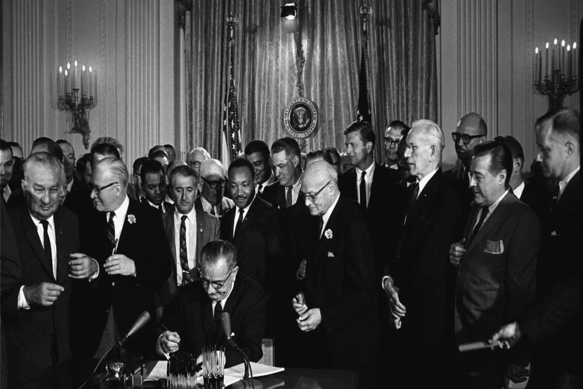 Black and white photo of a group of men and women standing or sitting in front of a building.
