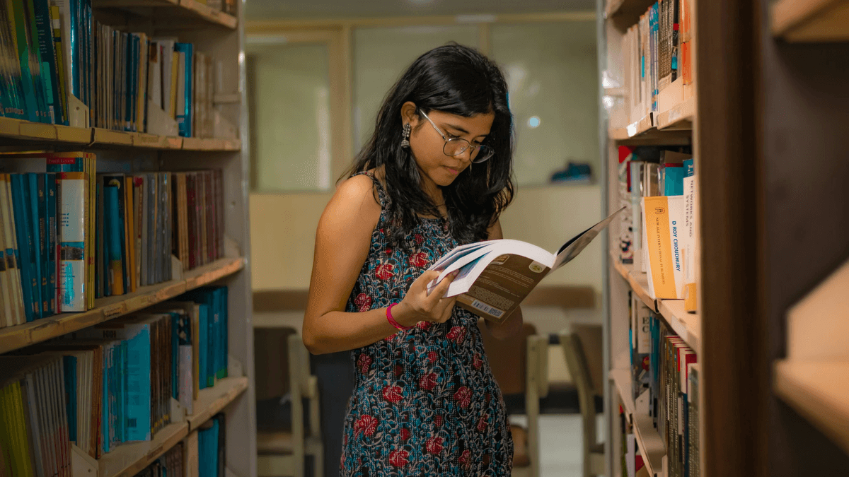 Oriental female wearing glasses and flower-patterned dress while standing in a library aisle reading a book. 