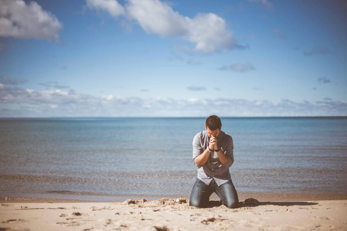 Image of white male with clasped hands kneeling down with by body of water.
